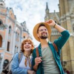 Mladý pár usmiatych turistov objavuje historické mesto s krásnou architektúrou. Muž nosí slamený klobúk a okuliare, žena má šatku okolo krku. Na pozadí sú historické budovy s detailnou fasádou a vežou.