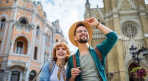 Mladý pár usmiatych turistov objavuje historické mesto s krásnou architektúrou. Muž nosí slamený klobúk a okuliare, žena má šatku okolo krku. Na pozadí sú historické budovy s detailnou fasádou a vežou.